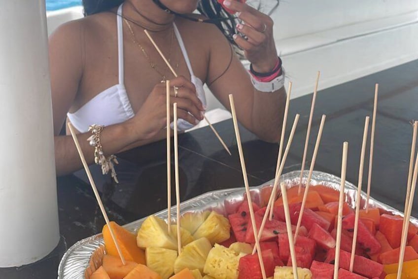 Una persona in costume bianco che tiene un drink, con un vassoio di spiedini di frutta davanti. 5