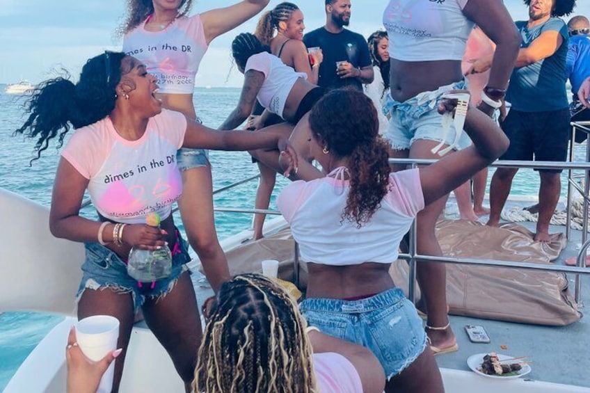 Un gruppo di persone su una barca, alcune vestite con abiti estivi leggeri, con un corpo d'acqua e un cielo sereno sullo sfondo. 4