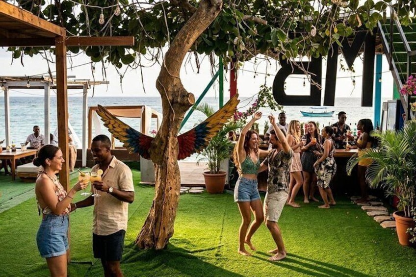 Tropische buitenomgeving met een houten pergola, een boom versierd met kleurrijke decoraties, en uitzicht op de zee. 1