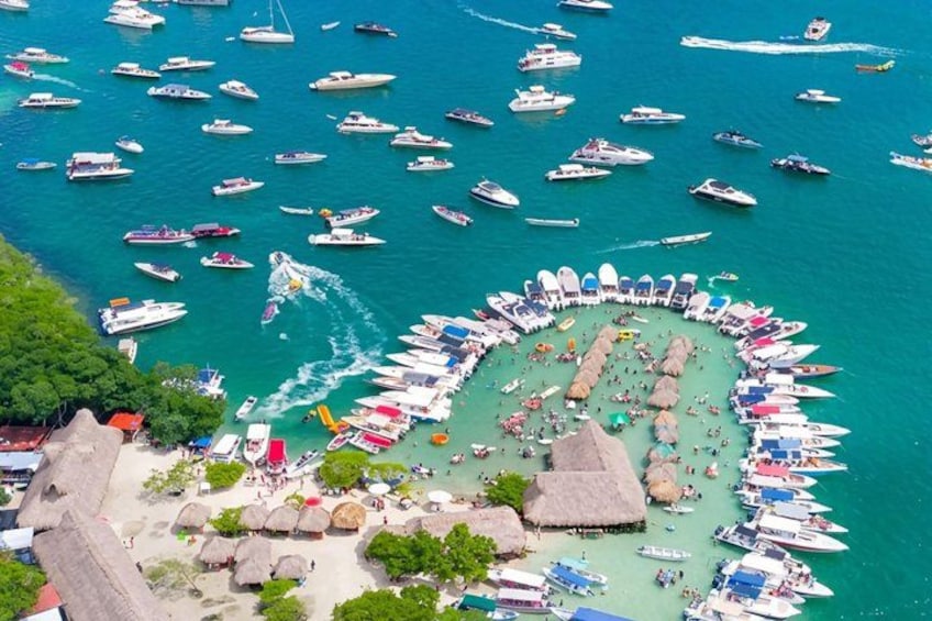 Un puerto deportivo con numerosas embarcaciones amarradas en forma circular, rodeado de una playa arenosa con cabañas de techo de paja y sombrillas. 4