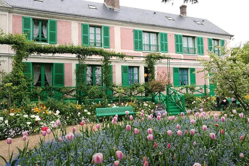Ein rosafarbenes und weißes Gebäude mit grünen Fensterläden, ein Garten mit Tulpen und eine grüne Bank. 1