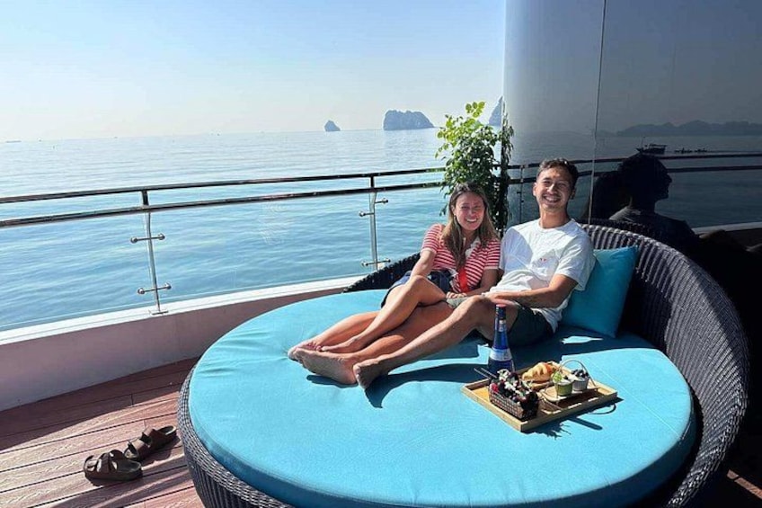 Deux personnes sont assises sur une terrasse extérieure bleue, dotée de coussins, d’où l’on peut admirer la mer et des îles lointaines. 3