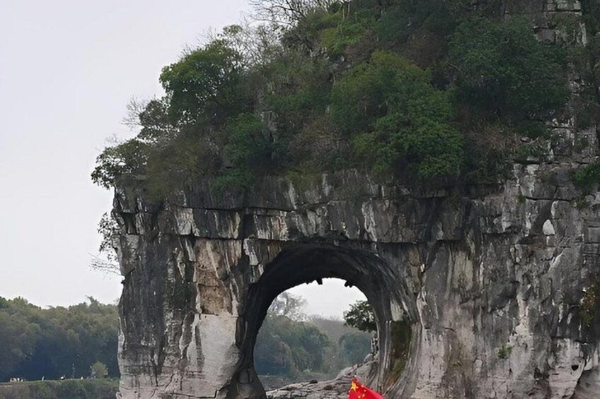 A large rock arch bridge with greenery on top and a red flag at the base. 2