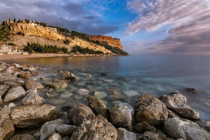 A rocky shoreline with clear water, a cliff with buildings, and a sunset sky. 3