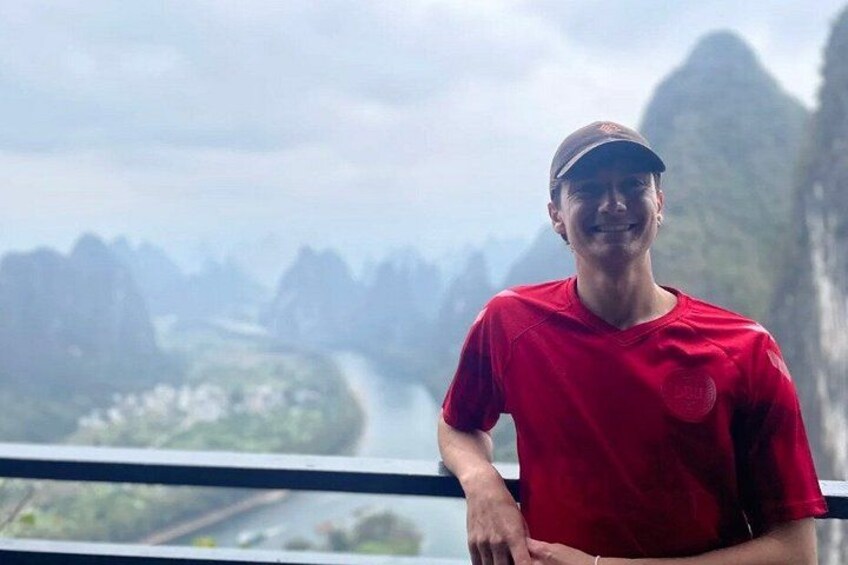 Une personne portant un t-shirt rouge, debout près d’une rambarde, avec un paysage montagneux en arrière-plan. 3