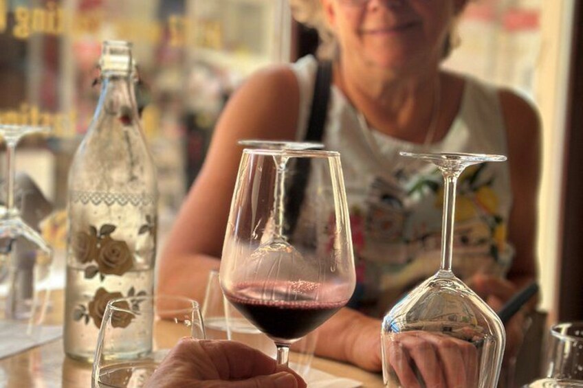 Una persona sostiene una copa de vino con líquido rojo, una botella transparente con motivos florales y otra copa de vino sobre una mesa. 4