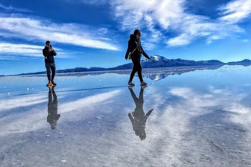 Two people walking on a reflective frozen lake with mountains in the background. 3