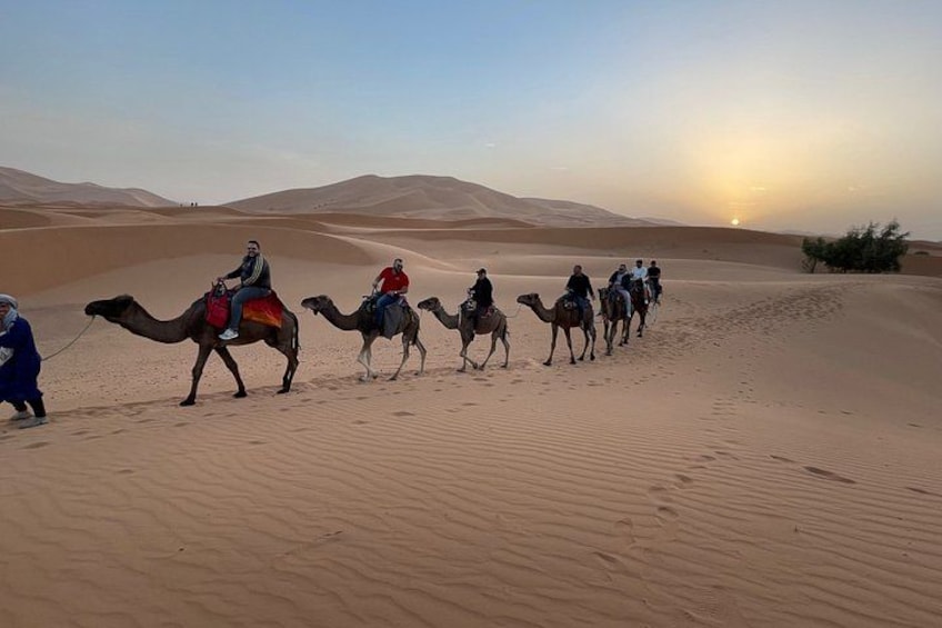 A group of people riding camels across a desert landscape with sand dunes and a sunset in the background. 1