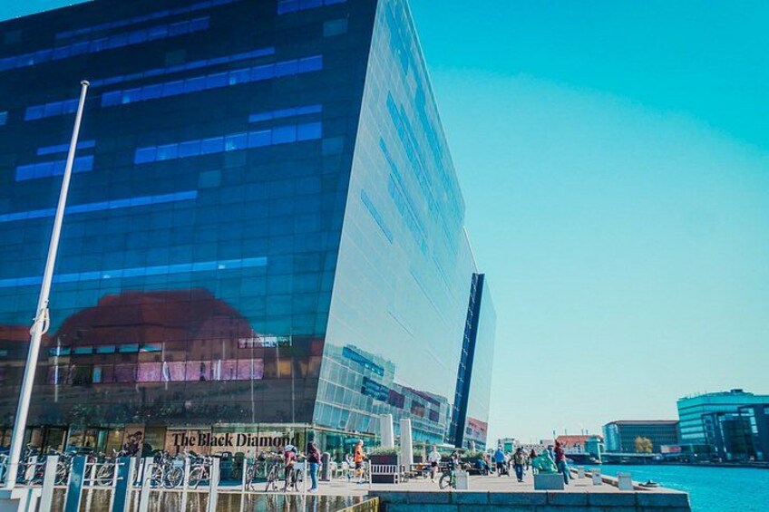 En moderne glasbygning med refleksioner, en klar blå himmel og et livligt havneområde med mennesker og cykler. 3