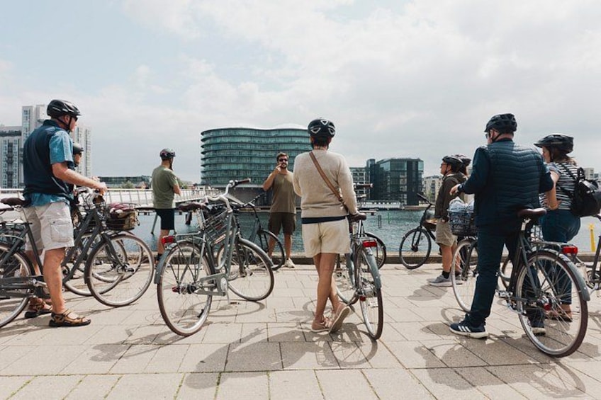 Personer står med sina cyklar nära en hamn, med byggnader i bakgrunden. 1