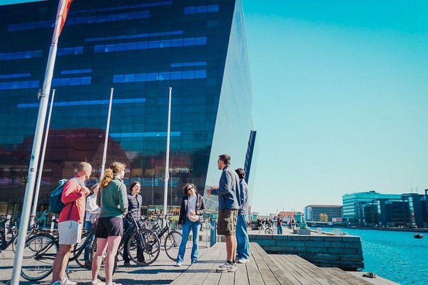 Personer står på en träbrygga vid ett vattendrag, med cyklar och en modern byggnad i bakgrunden. 2
