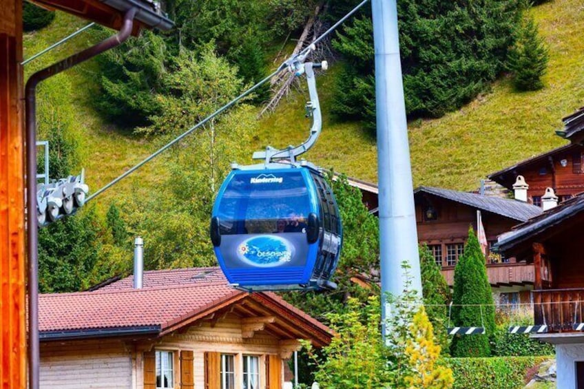 En blå linbana med texten ”Bergbahn”, en träbyggnad med ett rött tegeltak och en sluttning täckt av grönska. 2