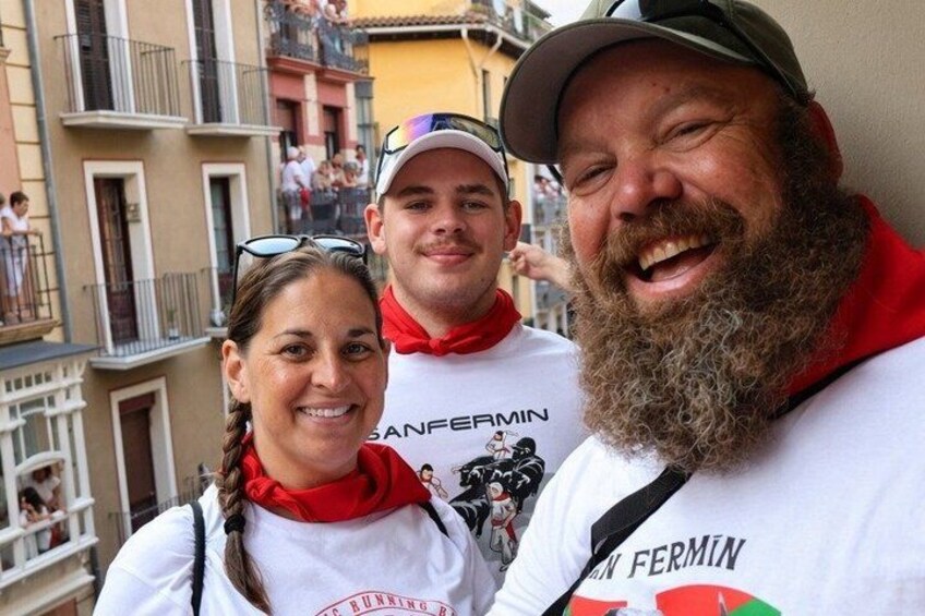 Une personne portant une chemise blanche, un foulard rouge et arborant un logo, se tient dans un quartier urbain, avec des immeubles et des balcons en arrière-plan. 2