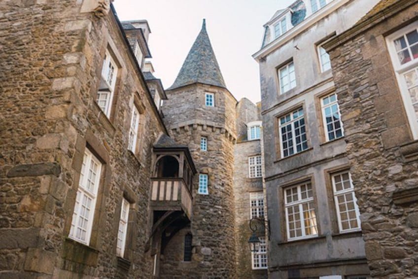 A narrow medieval street with stone buildings, a turreted tower, and a wooden balcony. 5