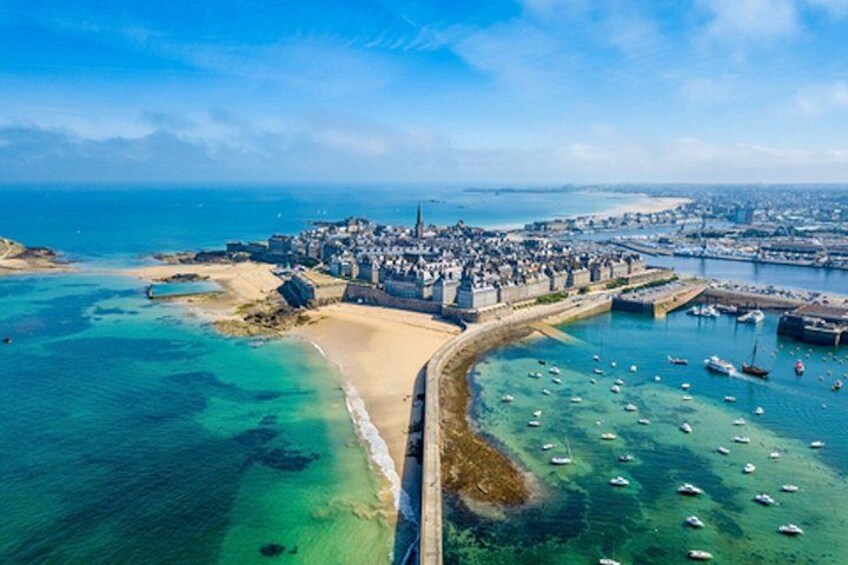 An aerial view of a coastal town with a sandy beach, a fortified structure, and a harbor with boats. 4