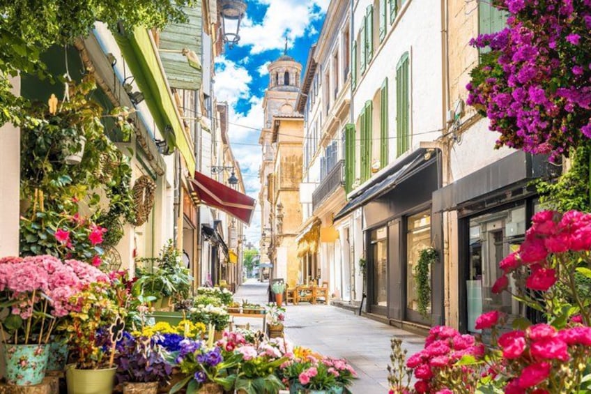 A narrow street with colorful flowers, buildings with shutters, and a bell tower in the distance. 1