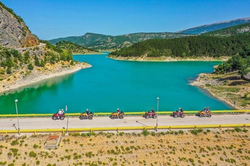 Un lago turquesa rodeado de colinas, un camino pavimentado con cuatro vehículos todo terreno, y una barrera de seguridad amarilla. 1