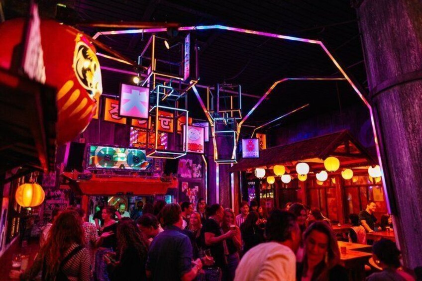 A crowded indoor venue with neon lights, a DJ booth, and people socializing. 1