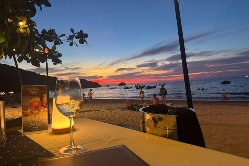 A beachside table with a glass of wine, a menu, and a pot, overlooking the sea with boats and a sunset. 5