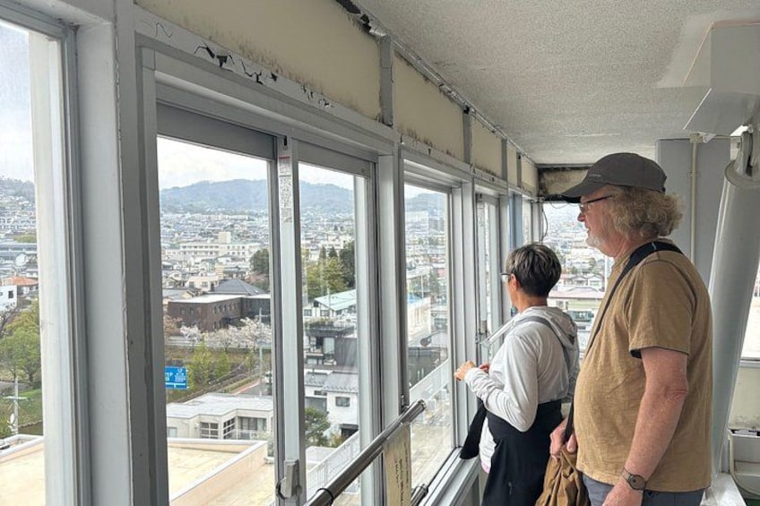 Two people standing by a large window overlooking a cityscape with buildings and a distant mountain. 2