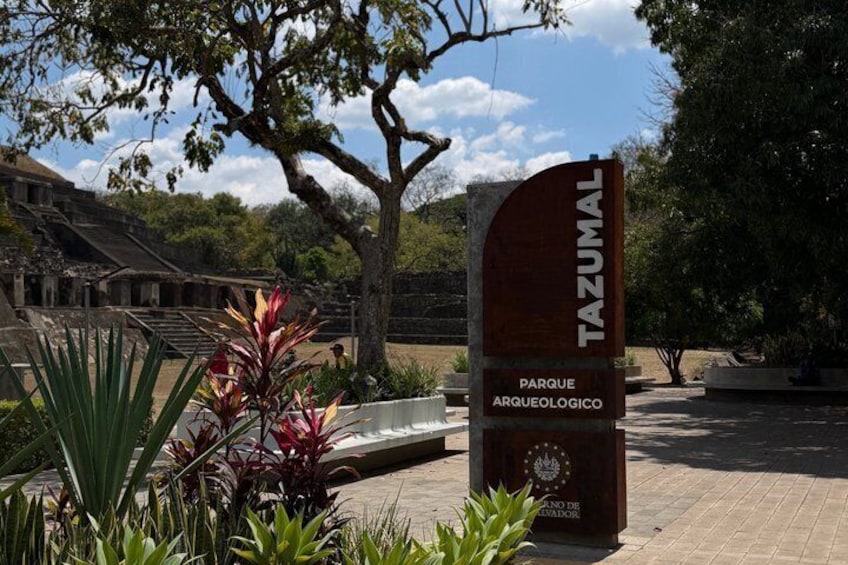 A sign for 'Tazumal Parque Arqueológico' with a stone staircase and surrounding greenery. 1