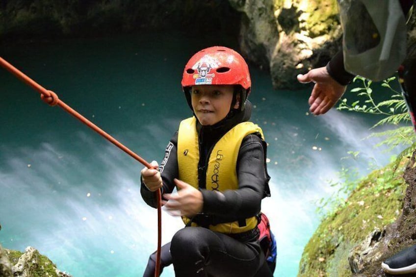 A person in a yellow life jacket and red helmet, holding a rope, near a body of water with a rocky background. 5