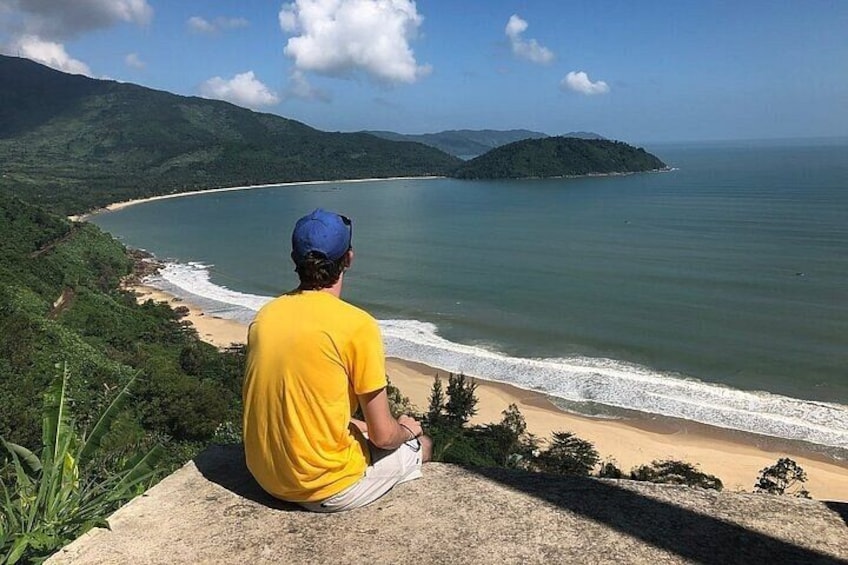 A person sitting on a ledge overlooking a beach with waves, greenery, and distant hills. 5