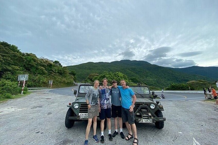 Four people standing in front of a military vehicle on a road with a mountainous landscape in the background. 3