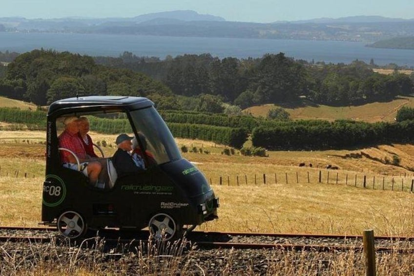 A small black car with a canopy, labeled 'railCruiser.com,' parked on a railway track with a scenic view of hills and a body of water in the background. 2
