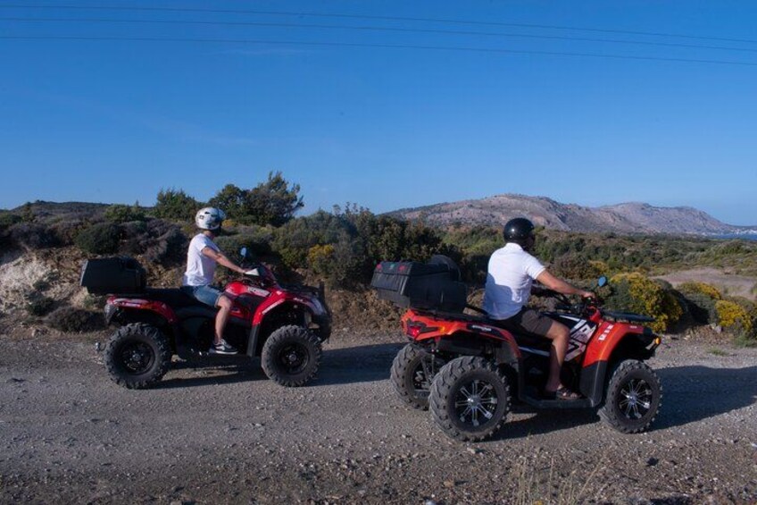 Two people riding red ATVs on a gravel road with a mountainous landscape in the background. 1