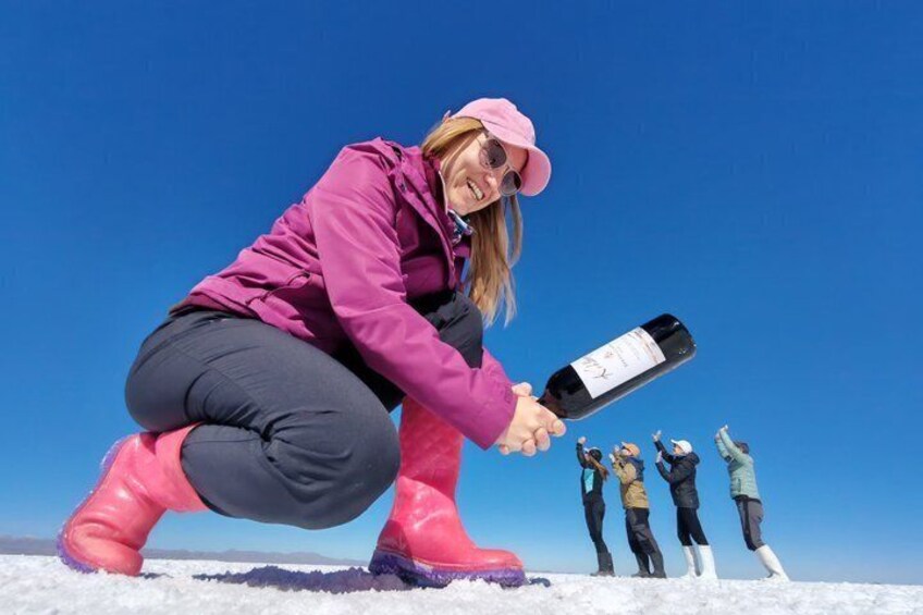 A person in pink boots and a pink jacket holding a bottle of wine, with a clear blue sky in the background. 5