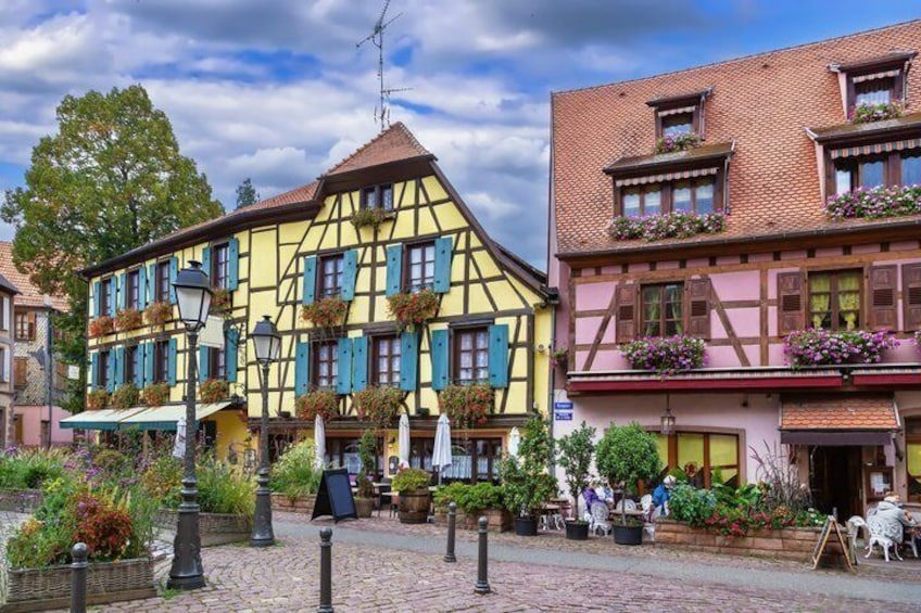 A cobblestone street with colorful half-timbered buildings, outdoor seating, and potted plants. 1