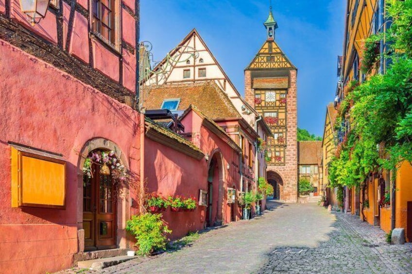A cobblestone street lined with colorful half-timbered buildings, a tower with a clock, and lush greenery. 3