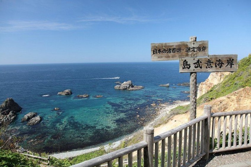 Vista costera con un cartel de madera, mar azul cristalino, afloramientos rocosos y una cerca de madera. 1