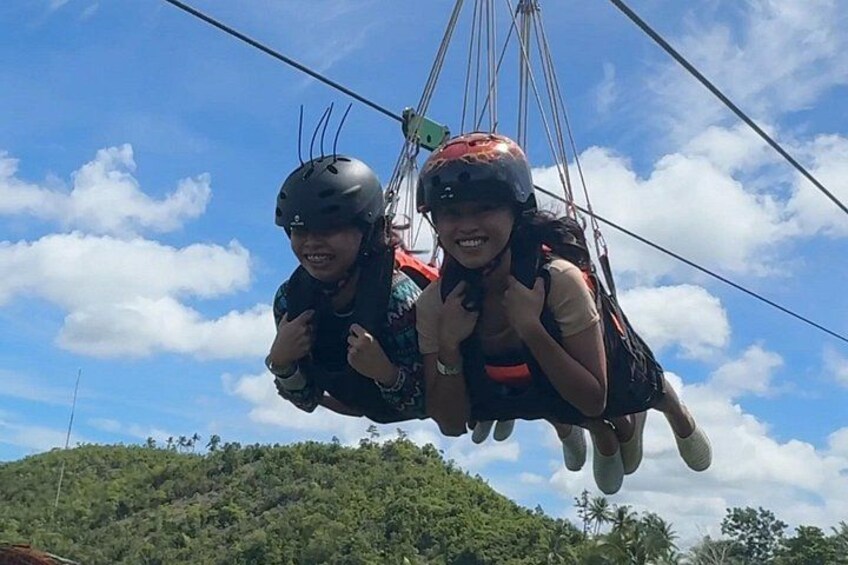 Two people suspended in mid-air by a cable, wearing helmets and harnesses, with a clear sky and a hilly landscape below. 4