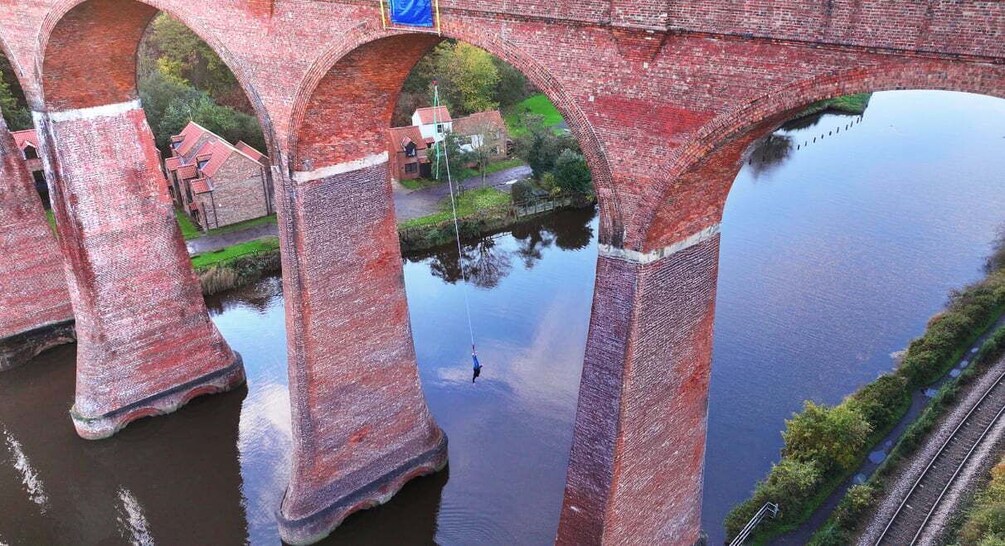 A red brick bridge with arches over a calm river, surrounded by greenery and a small building. 1