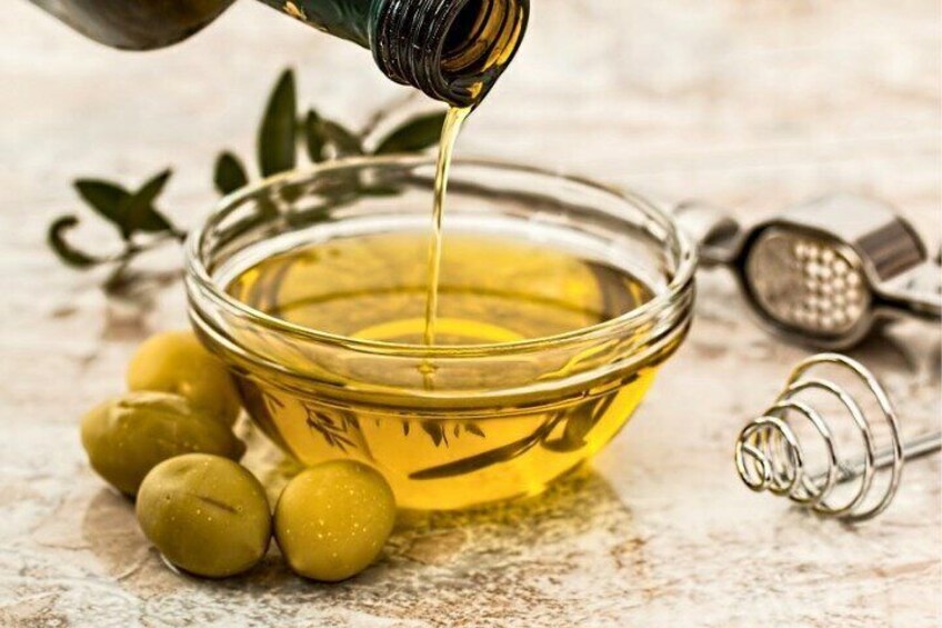 Olive oil being poured into a glass bowl, olives, and a grater on a wooden surface. 2