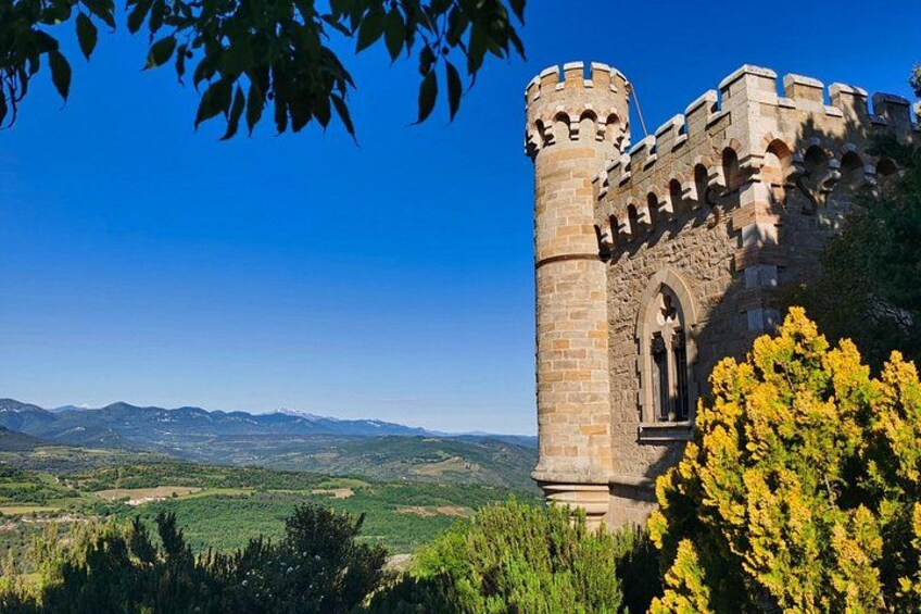 Un castillo medieval con una torre alta, rodeado de vegetación y montañas al fondo. 3