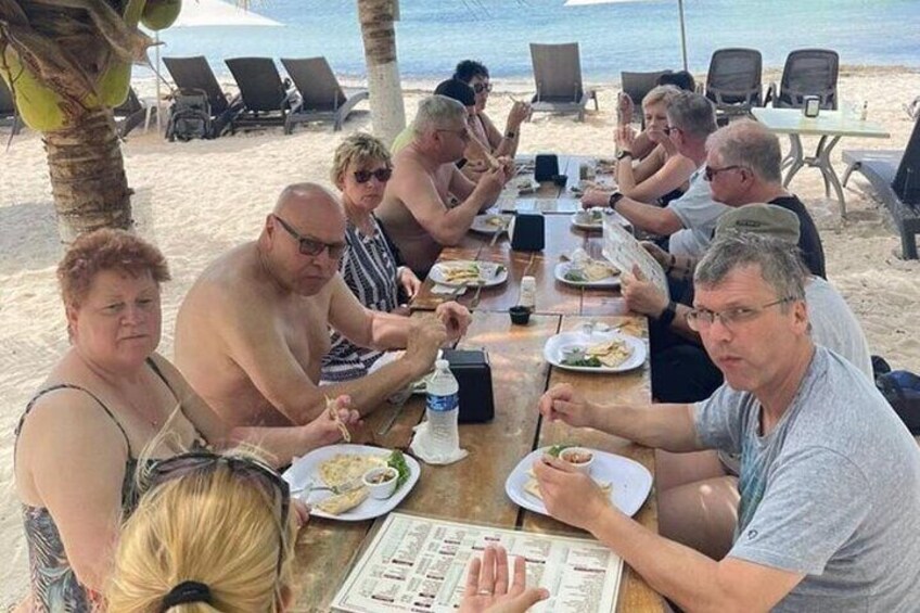 A group of people dining at a wooden table on a beach with palm trees and ocean in the background. 5