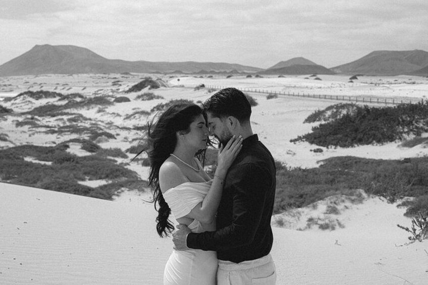 A couple embracing on a beach with sand dunes and a distant bridge. 1