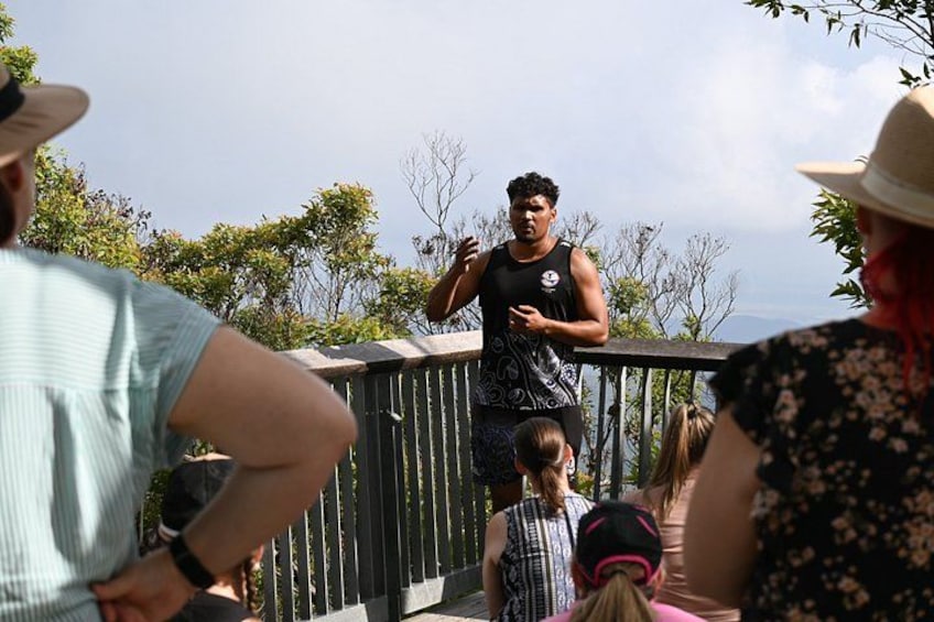 A person speaking to an audience on a bridge with trees in the background. 1