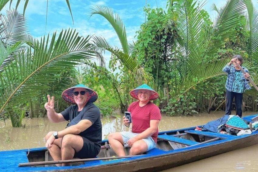 Two people in a boat, one wearing a hat, another holding a camera, surrounded by palm trees. 1