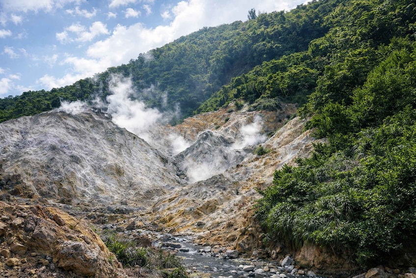 A geothermal landscape with steam vents, a rocky riverbed, and dense forested hills. 2