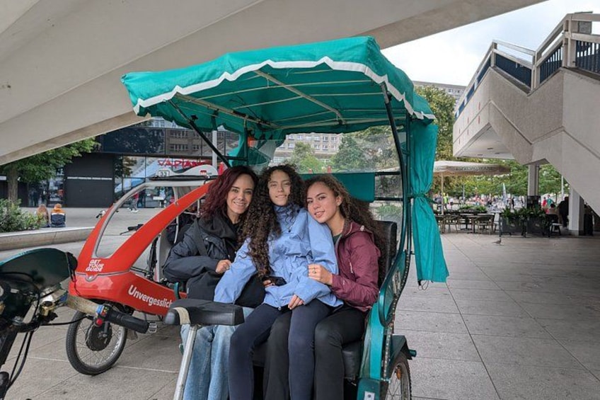 Trois personnes sont assises sur un vélo vert et rouge équipé d’un auvent, et celui-ci est garé sur un trottoir. 3