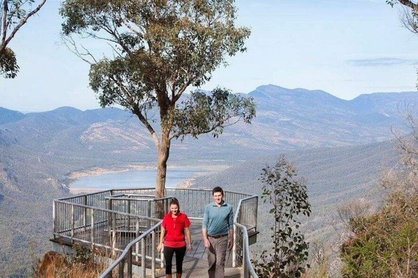 Two people walking on a metal bridge with a tree and a mountainous landscape in the background. 3