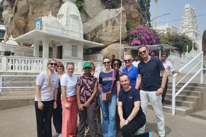 Een groep mensen poseert voor een tempel, met een grote rots en een trap op de achtergrond. 3