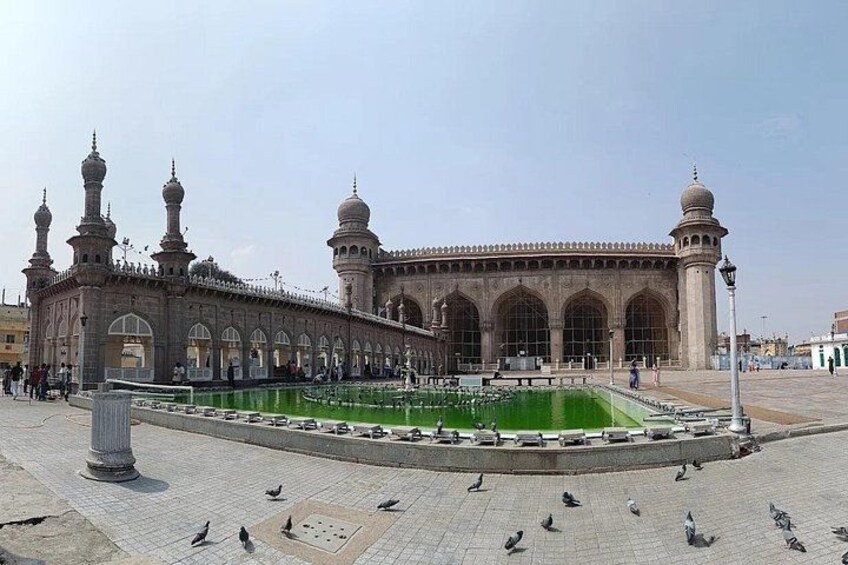 A large mosque with multiple domes and minarets, a central courtyard with a green pool, and pigeons on the ground. 2