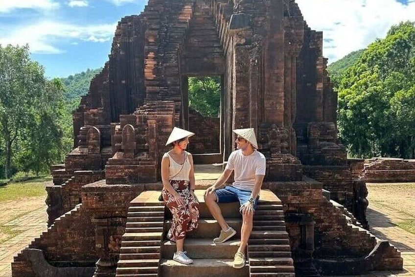 Twee personen zitten op oude stenen trappen voor een groot, gedeeltelijk vervallen tempel. 5