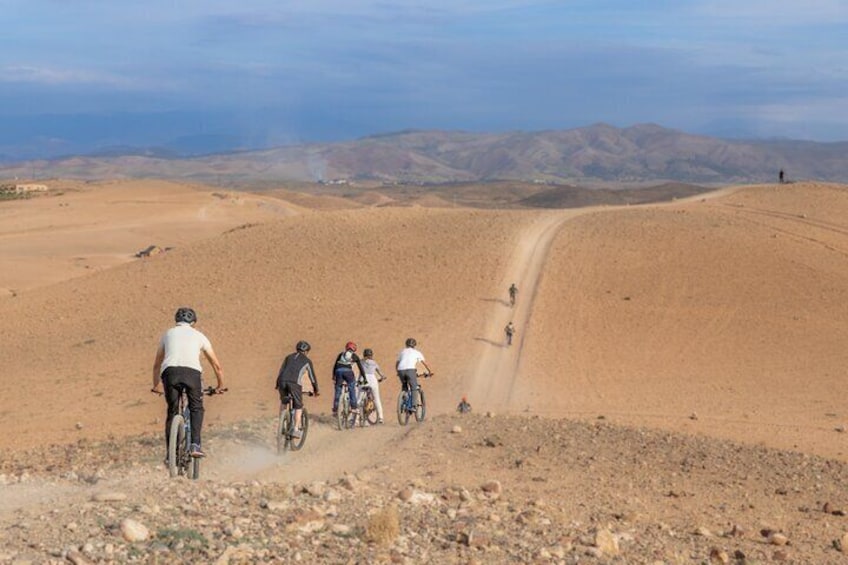 Un groupe de cyclistes sur un chemin de terre, dans un paysage désertique, avec des montagnes qui se dessinent à l'horizon. 3