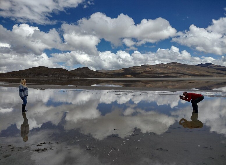 A reflective lake with mountains in the background and two people standing on the shore. 1